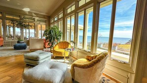 A spacious living room with large windows offering a view of the sea, a yellow armchair, a grey sofa, and a patterned rug. - The Chanler at Cliff Walk (Newport)