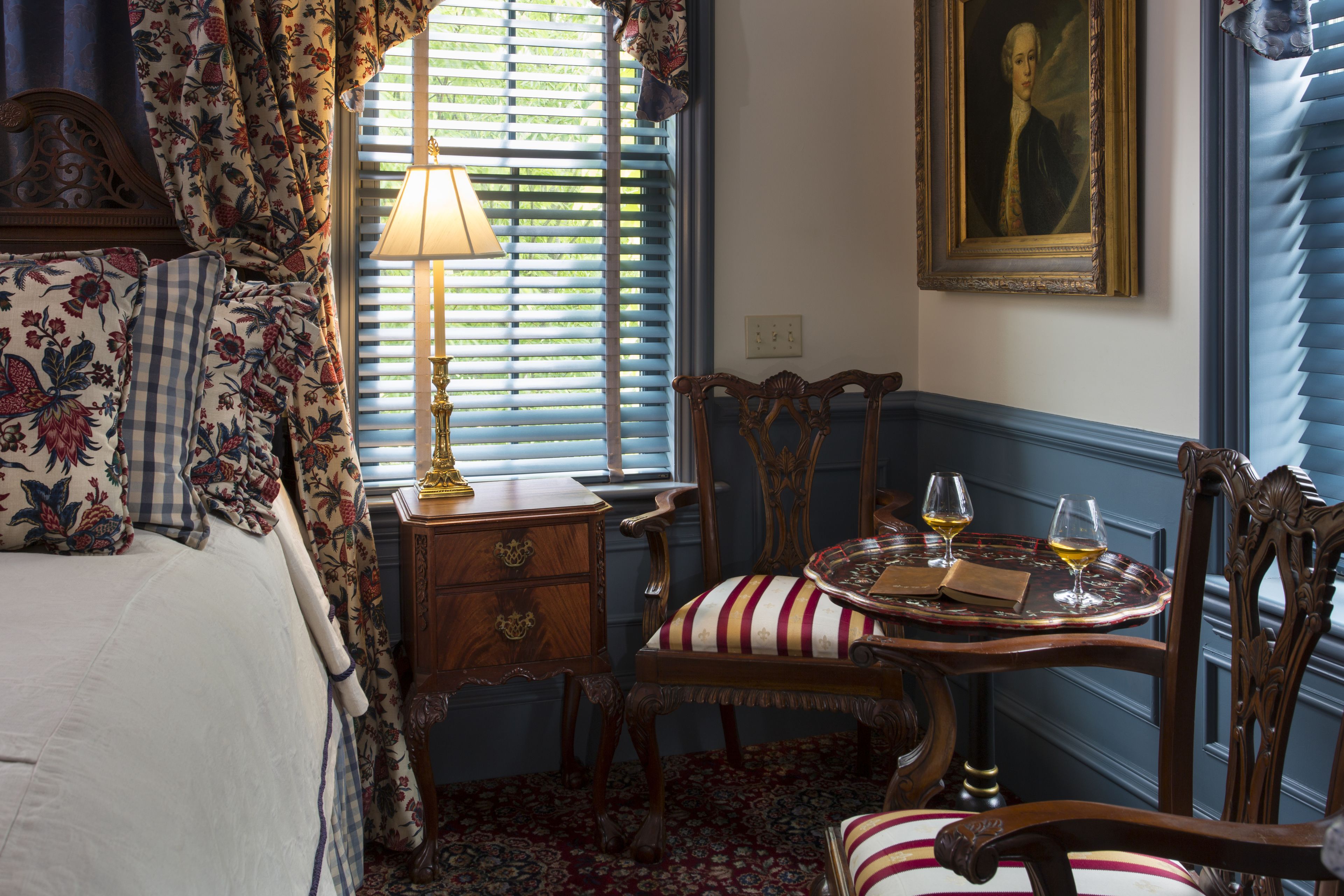 Une chambre à coucher avec un lit, une table de chevet, une lampe, une chaise, une table avec des verres et un tableau accroché au mur.
