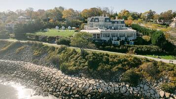 A large, multi-story house with a prominent balcony, situated on a hillside overlooking a rocky shoreline.
