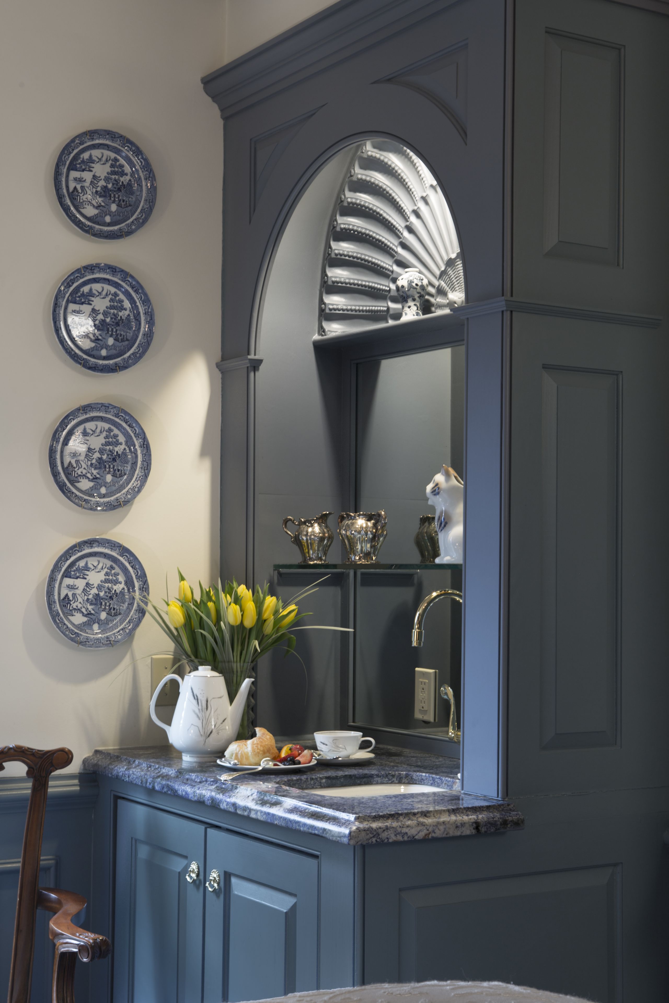 A kitchen with a marble countertop, a sink, and a mirror above it. There are blue and white plates on the wall and a vase with yellow flowers on the counter.