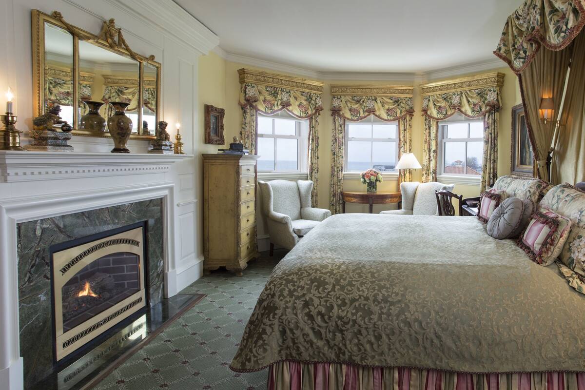 A bedroom with a fireplace, a large bed with ornate bedding, a sitting area with a chair and a table, and a view of the sea through the windows.