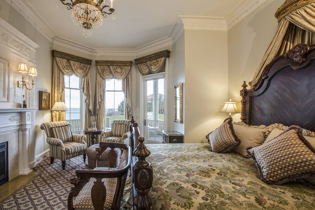 A bedroom with a large bed, two armchairs, a wooden side table, a fireplace, and a view of the outdoors through large windows.
