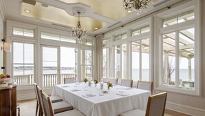 A dining room with a large table set for six, surrounded by chairs, large windows with a view of the sea, and a chandelier. - The Chanler at Cliff Walk (Newport)