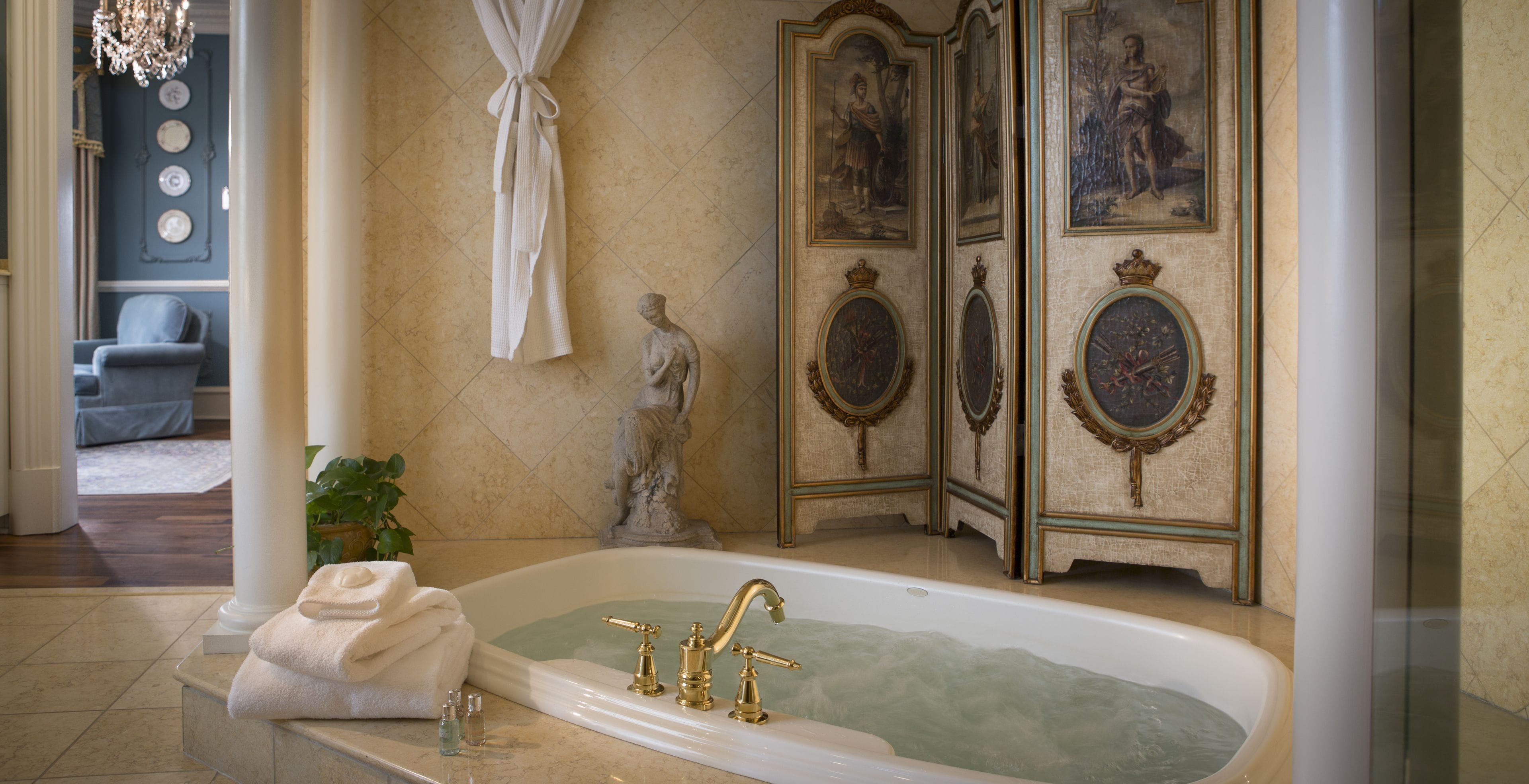 A luxurious bathroom with a freestanding bathtub, marble walls, and ornate decor including framed paintings and a statue.