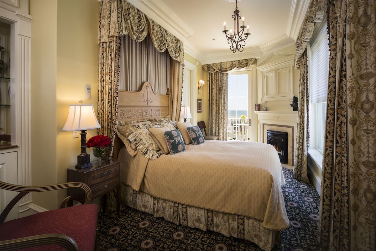 A traditional bedroom with a large bed, ornate curtains, a chandelier, and a fireplace.