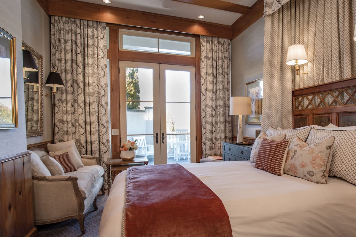 A bedroom with a large bed, a wooden headboard, two bedside tables with lamps, a chair, and a view through a glass door to an outdoor balcony.