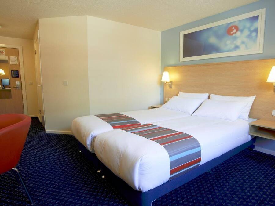 A hotel room with a bed, two bedside lamps, a red chair, and a painting on the wall.
