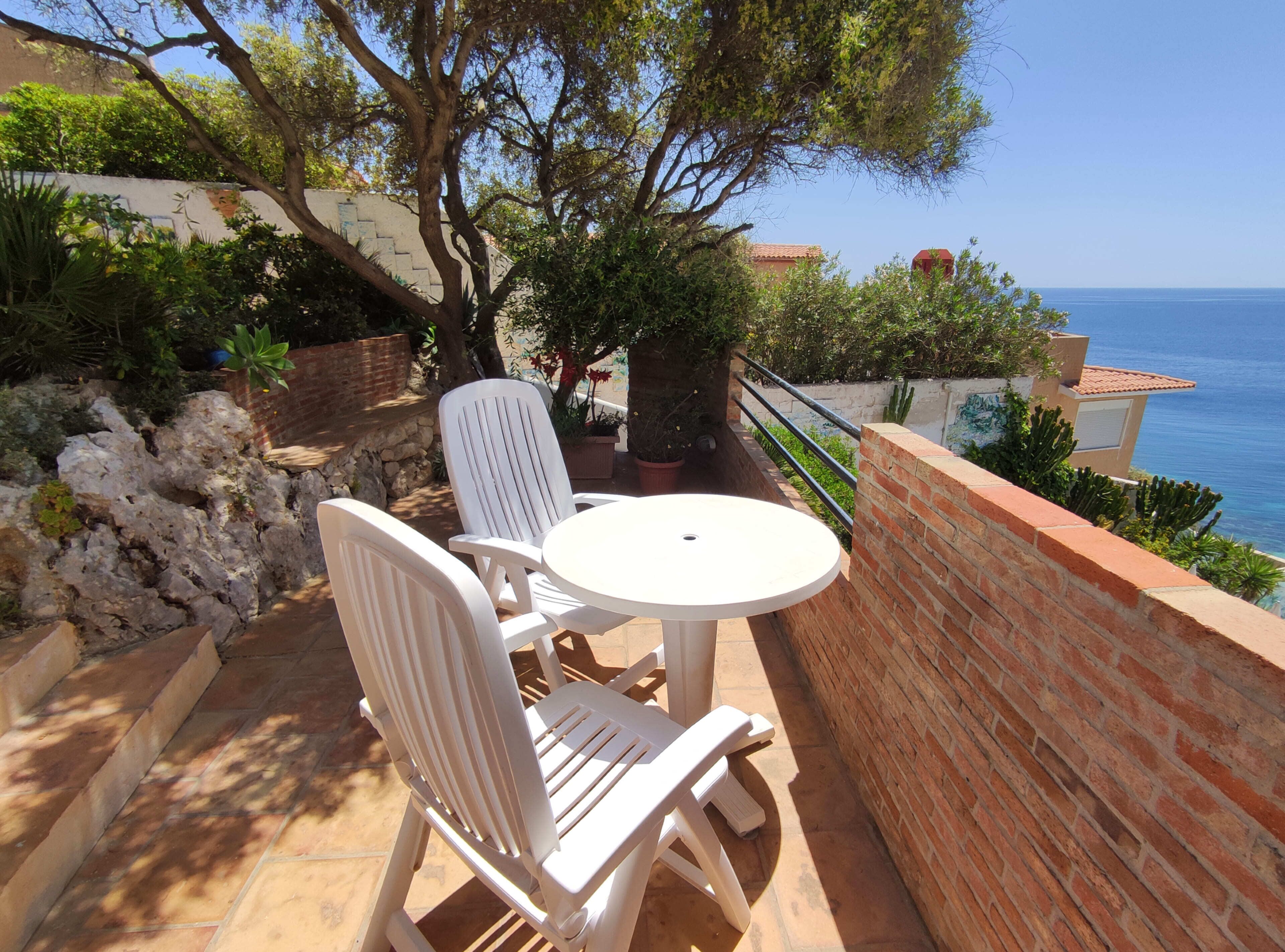 Une terrasse avec une table ronde et deux chaises blanches, offrant une vue sur la côte.