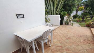 Un patio con una mesa y sillas blancas, rodeado de un jardín con diversas plantas y un cactus.