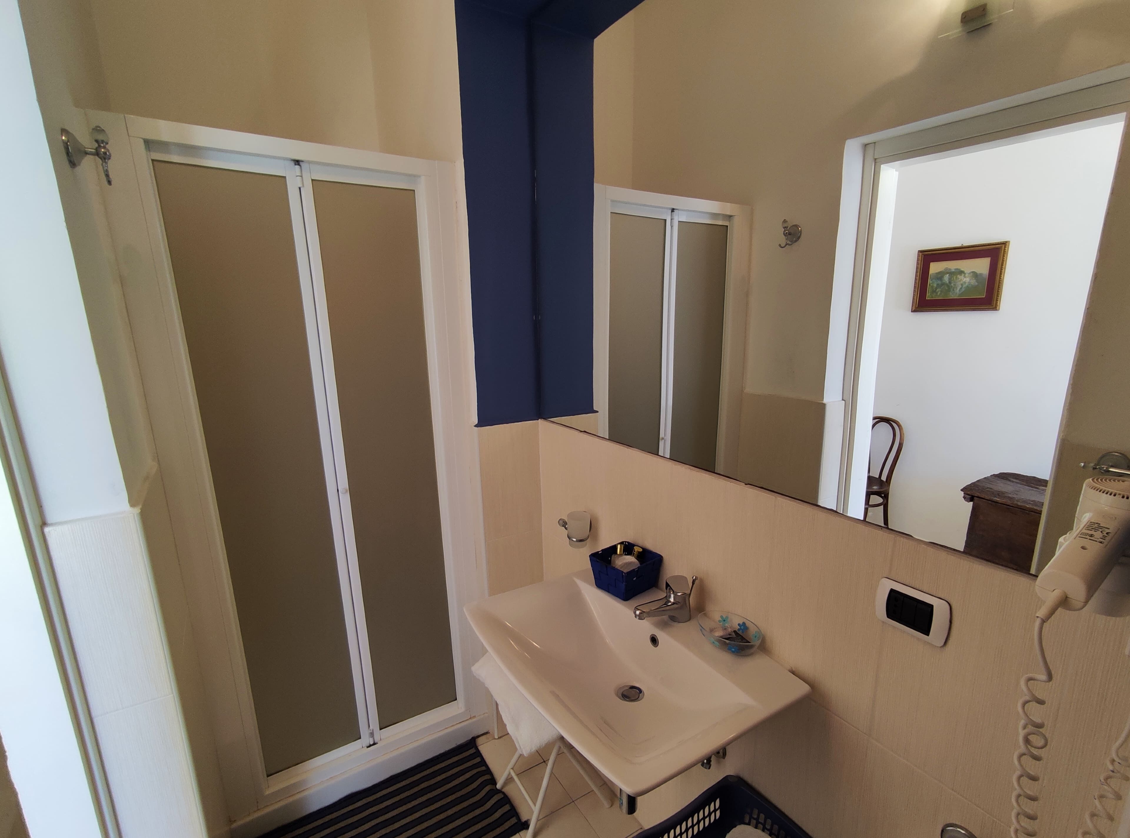 Baño con lavamanos blanco, espejo, cabina de ducha y cubo de basura pequeño azul.