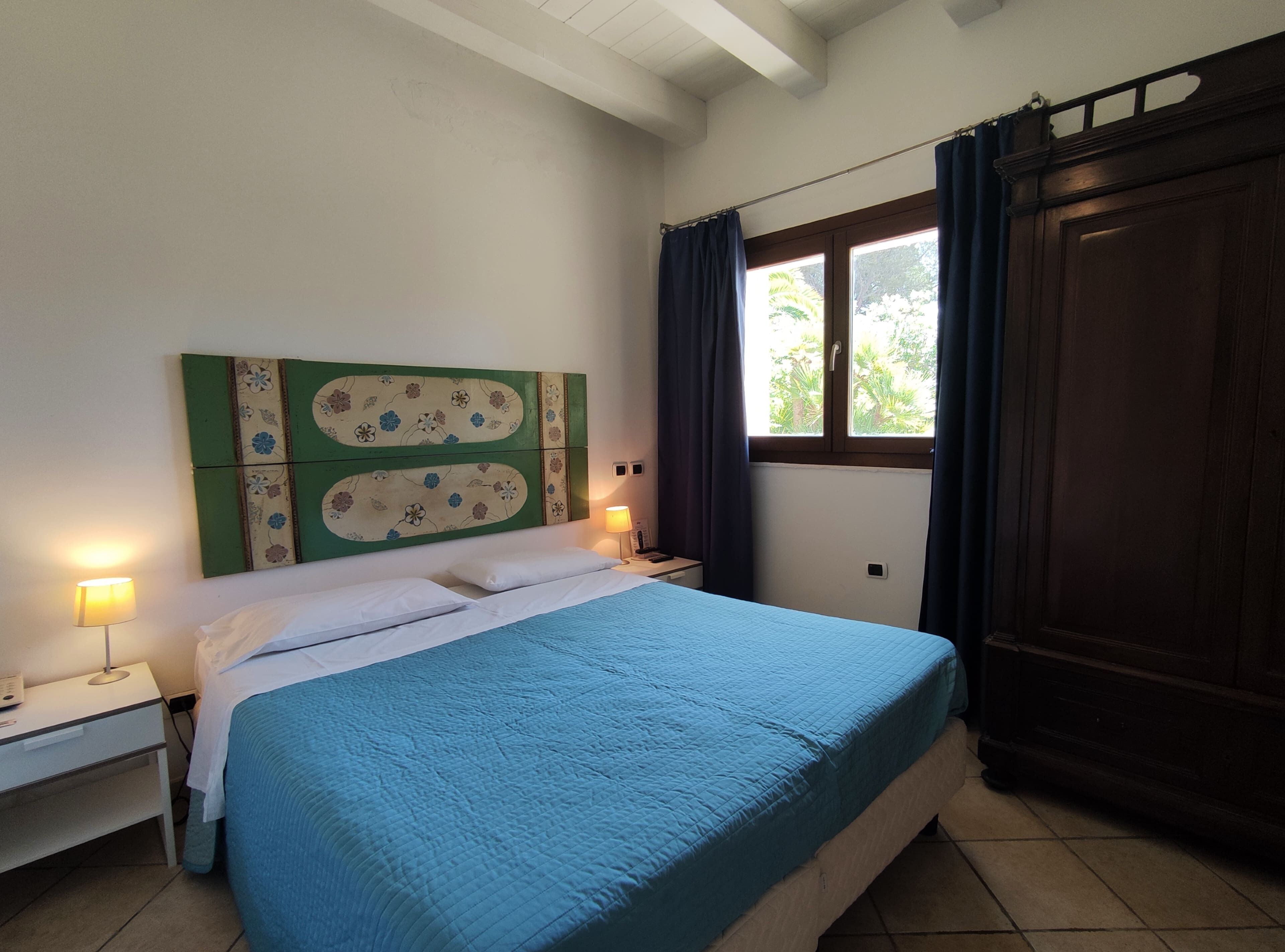 A bedroom with a bed, two bedside lamps, a nightstand, a window with curtains, and a wooden wardrobe.
