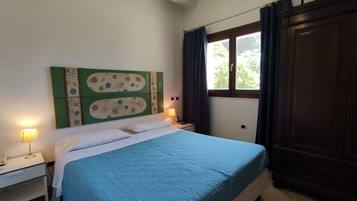 A bedroom with a bed, two bedside lamps, a nightstand, a window with curtains, and a wooden wardrobe.