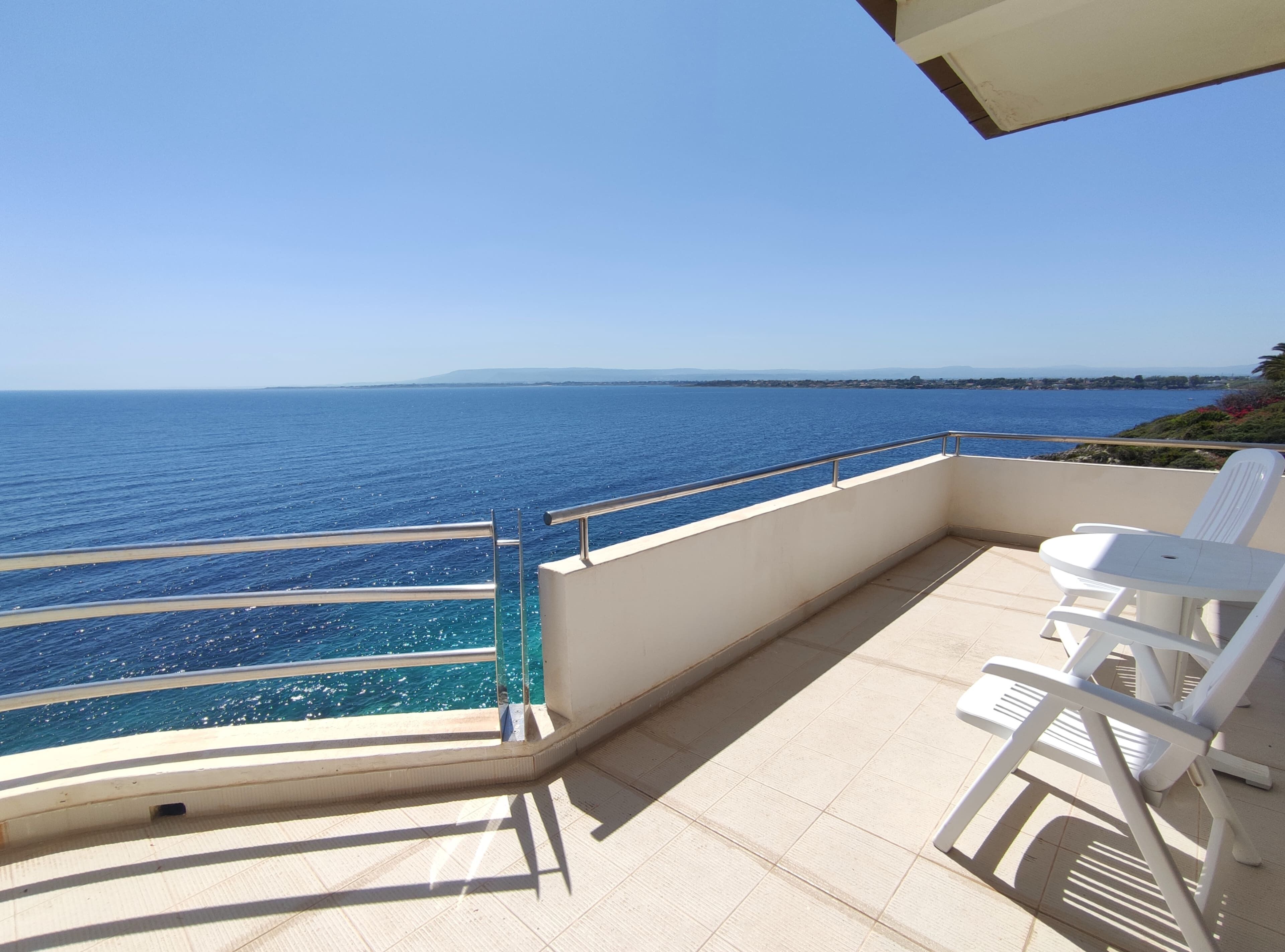 Un balcón con vistas al mar, una silla blanca y una mesita.