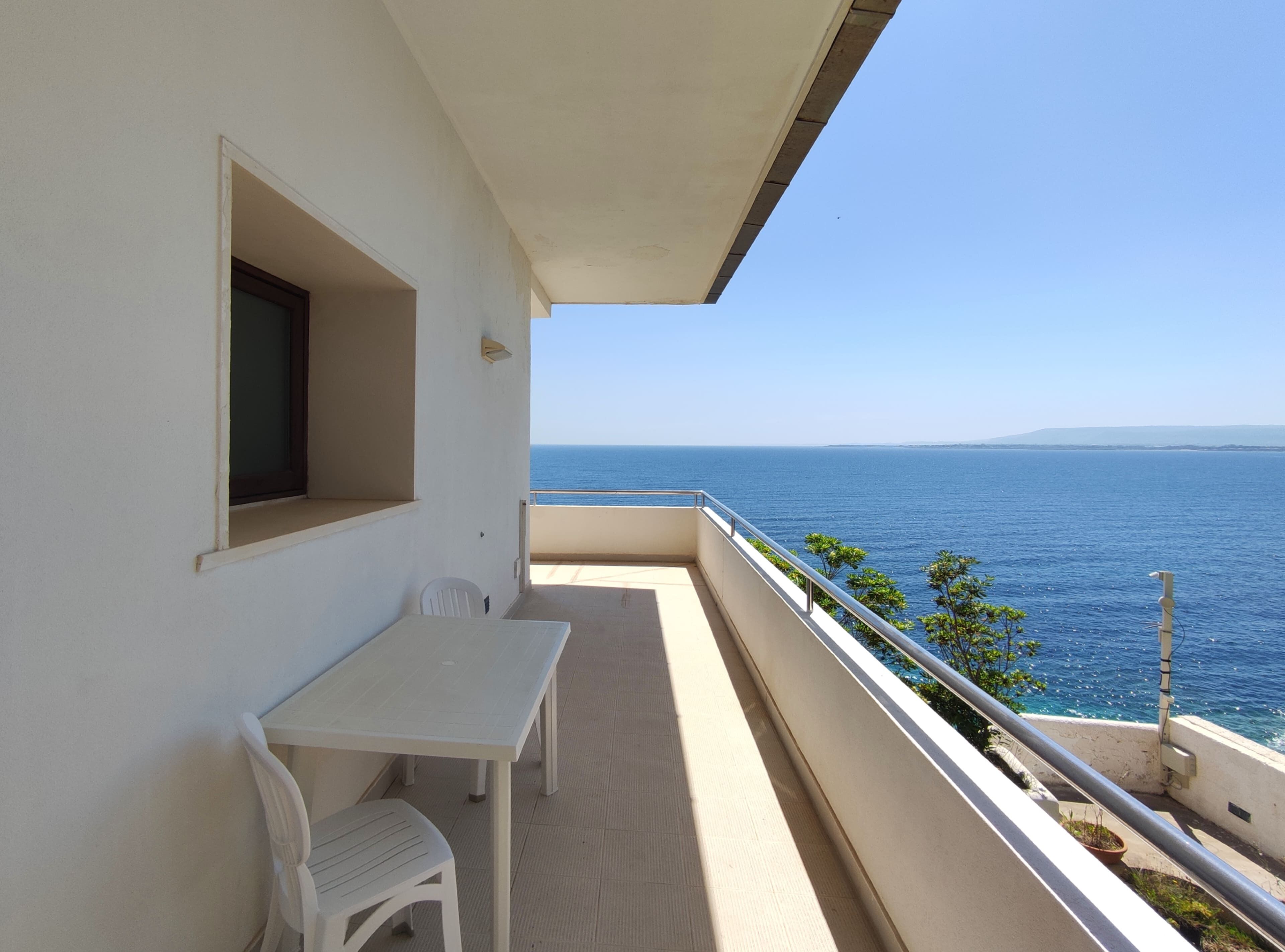 Un balcón con mesa y sillas, con vistas a un mar azul y una costa distante.