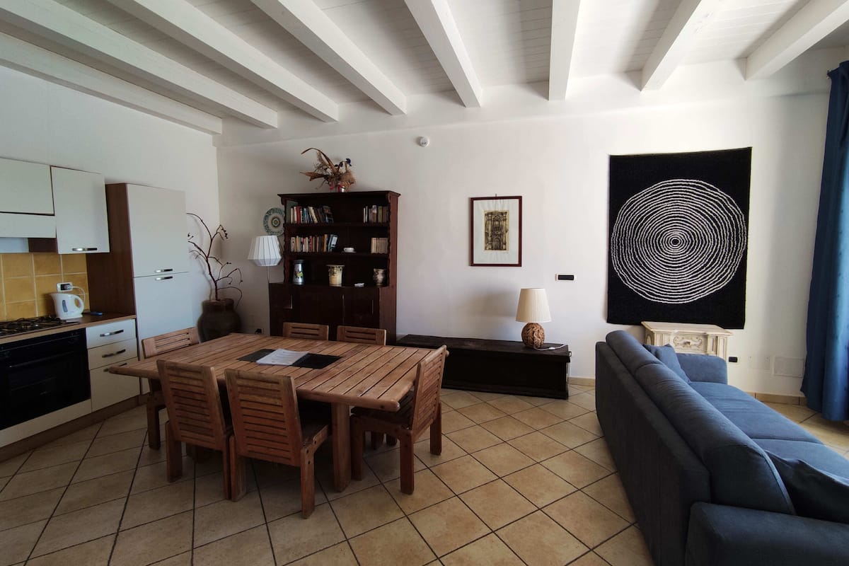 A living room with a dining table, chairs, a sofa, a bookshelf, and a kitchen area with cabinets and a stove.