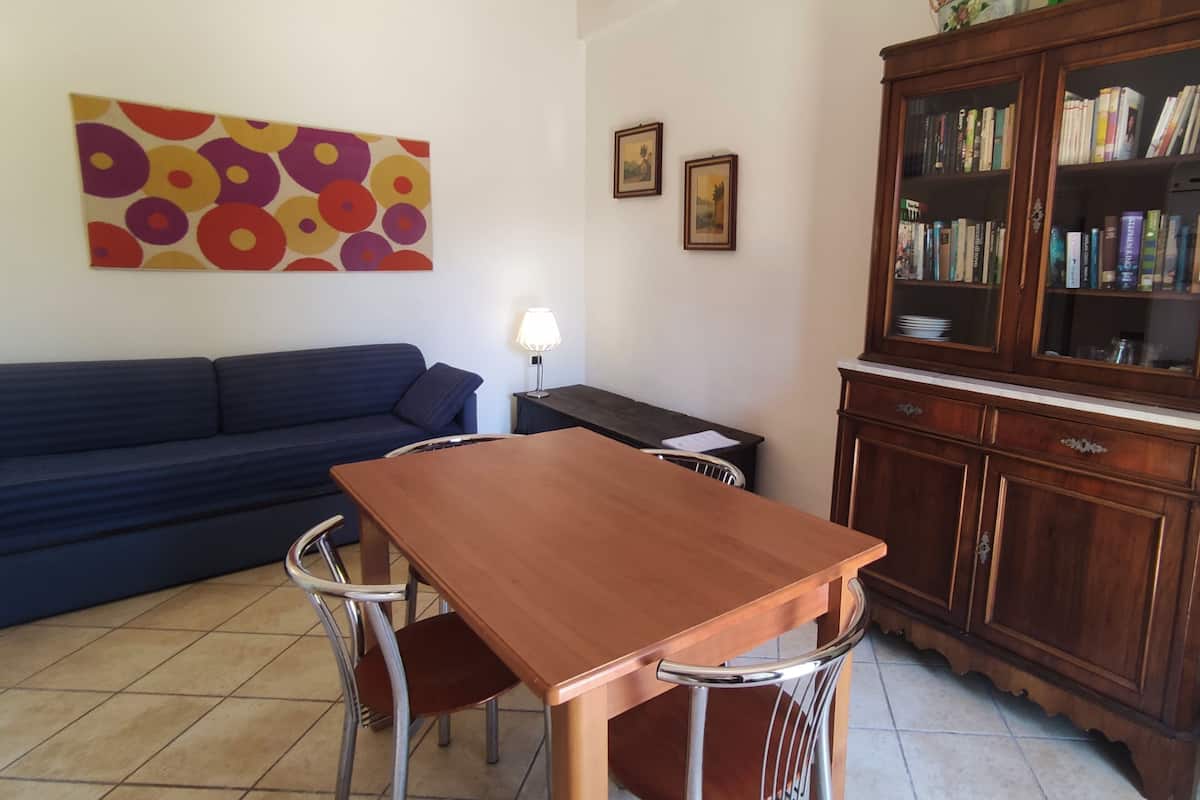 A living room with a sofa, a wooden dining table with chairs, a lamp, a bookshelf, and a colorful abstract painting.