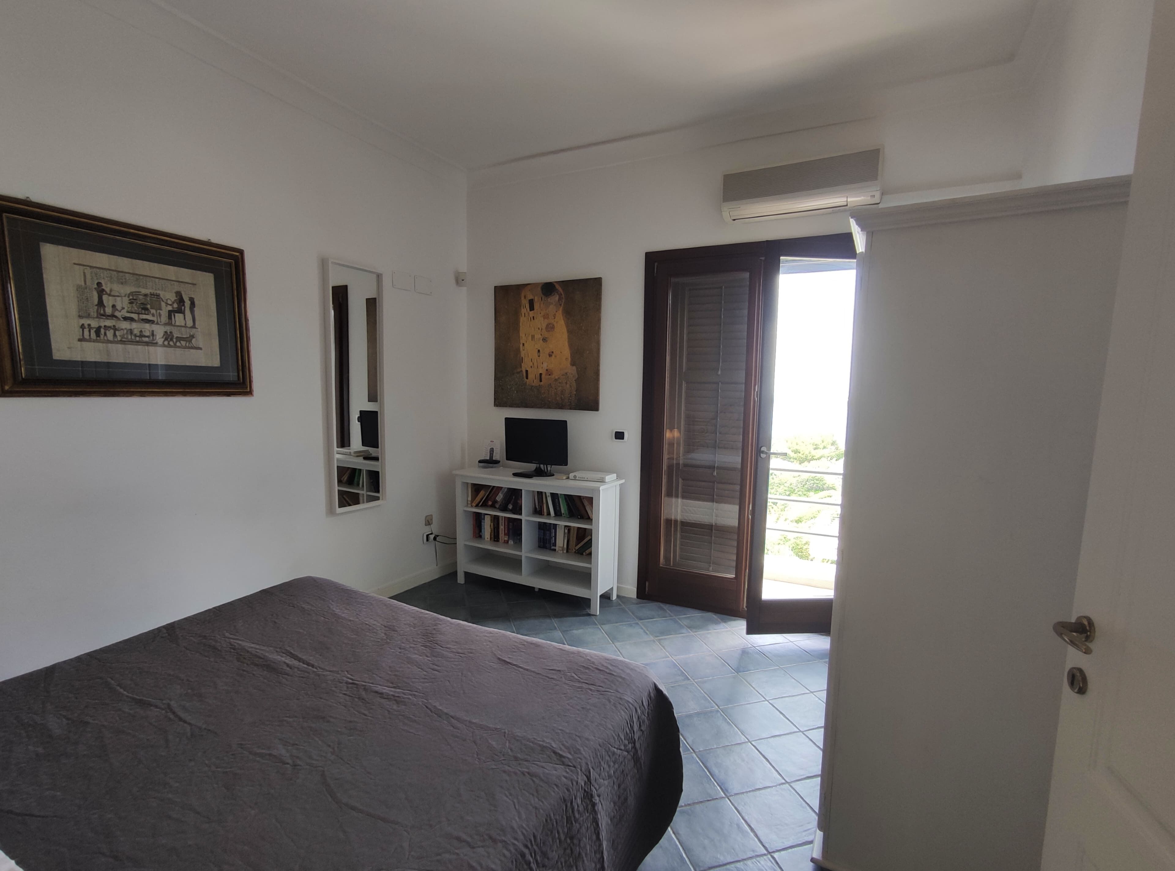 A bedroom with a bed, a TV on a white stand, a bookshelf, a framed picture on the wall, and a door leading to a balcony.