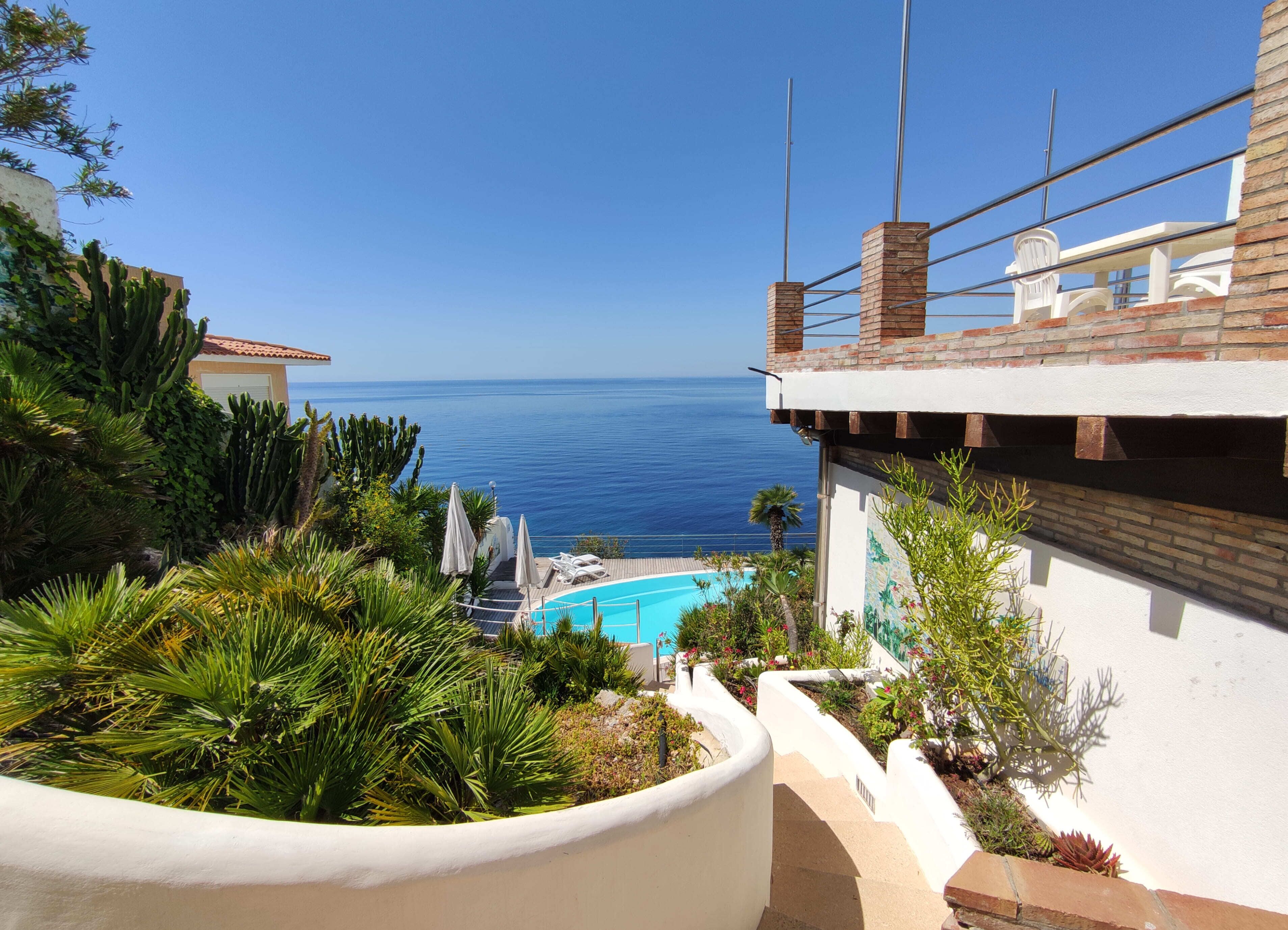 A pool with a view of the ocean, surrounded by greenery and a white building with a brick section.