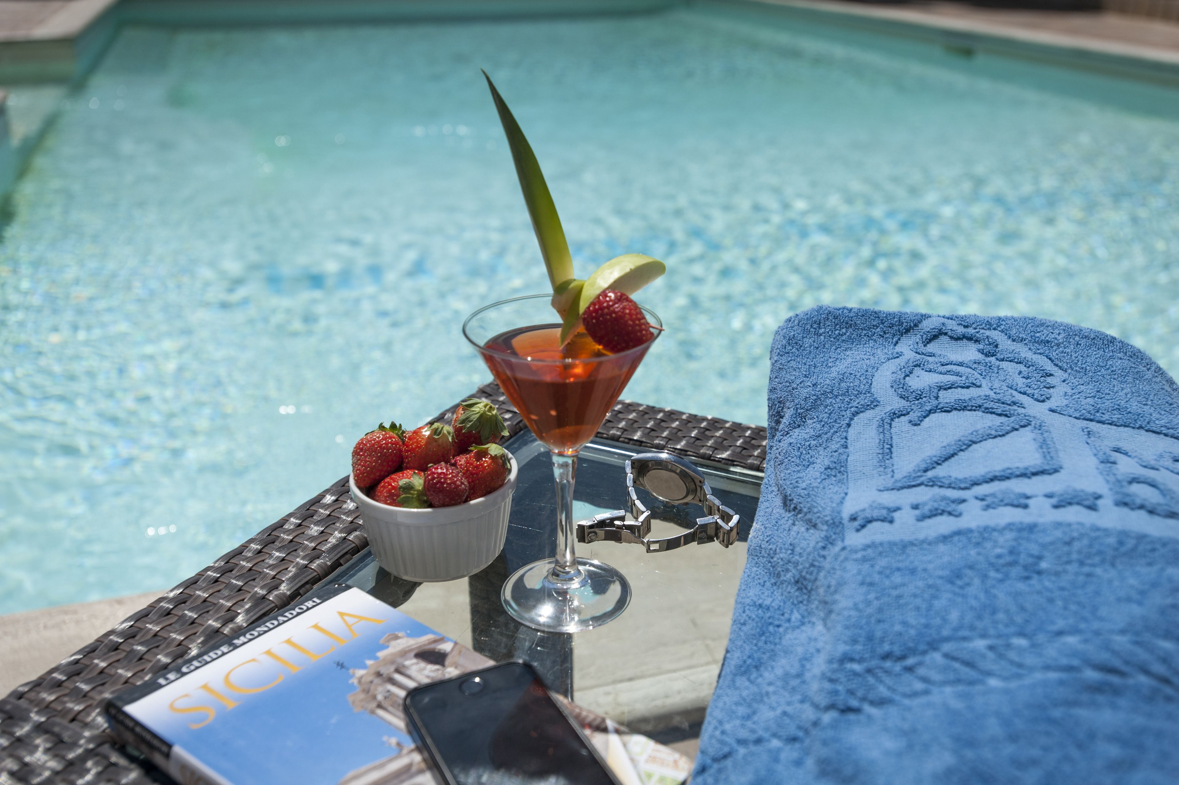 Une scène de bord de piscine avec un verre à martini, des fraises, un livre intitulé « Le Guide Mondodori Sicilia », un smartphone et une serviette ornée de monogrammes.