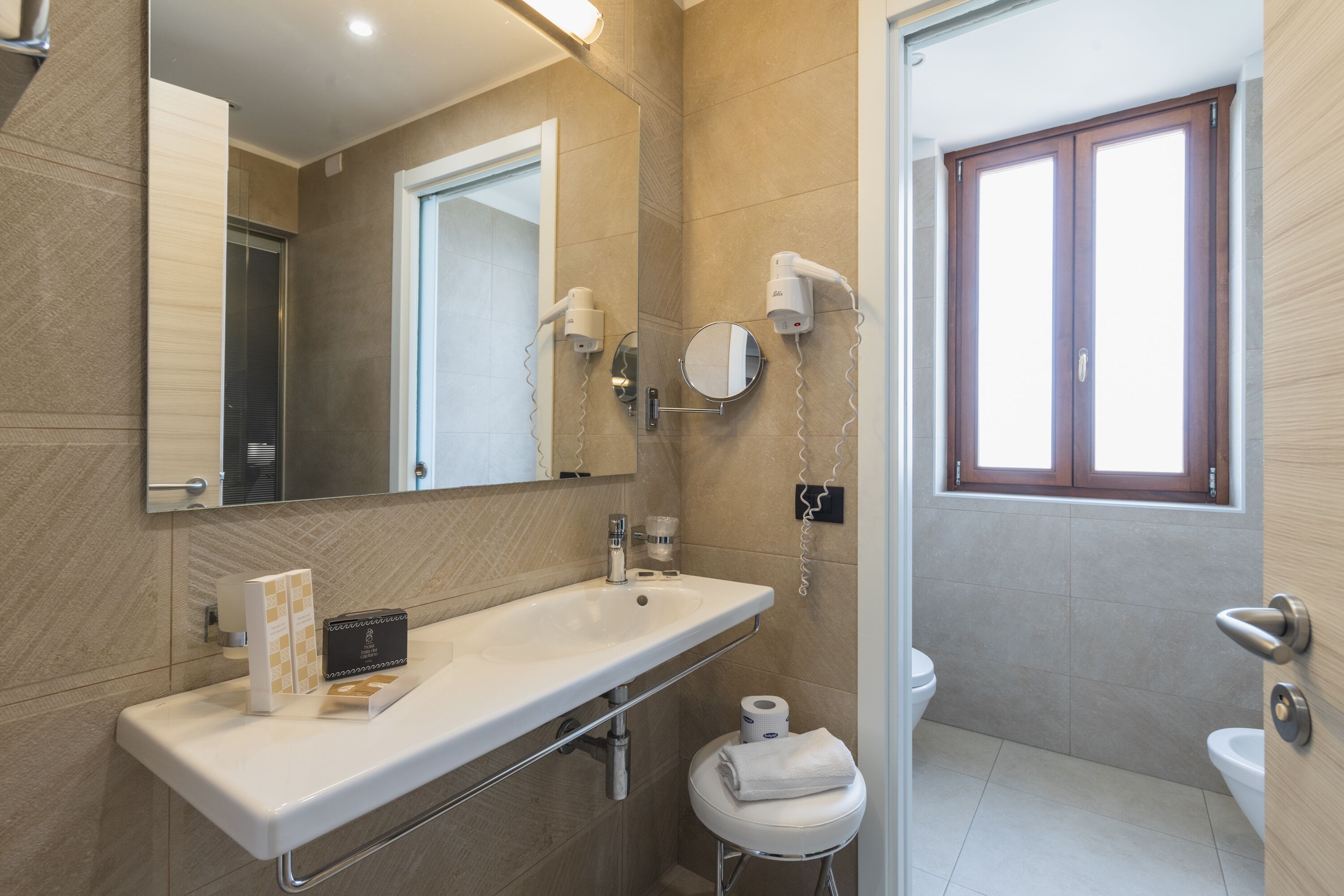 Un baño moderno con un espejo grande, un lavabo y un inodoro. Hay secadores de pelo montados en la pared y un taburete pequeño con una toalla.