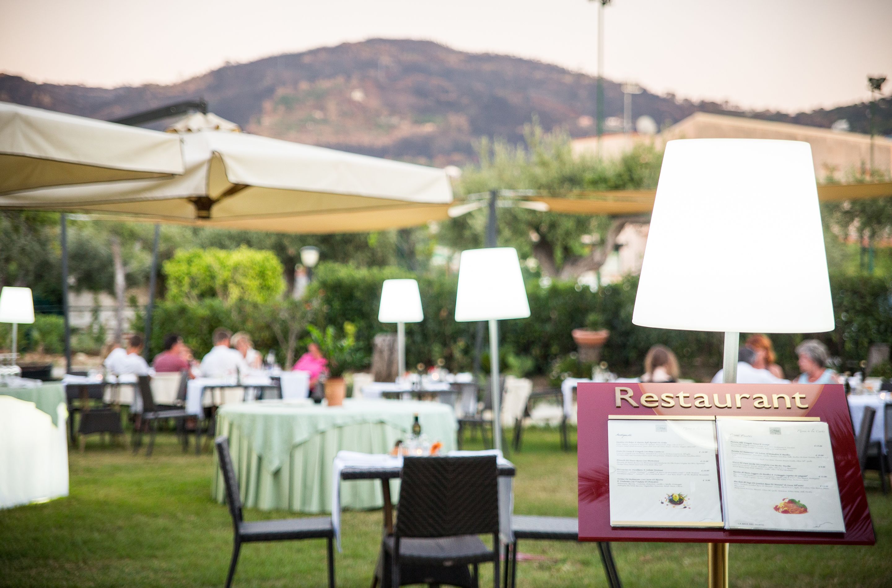 An outdoor restaurant with tables and chairs, a menu stand, and a mountainous backdrop.