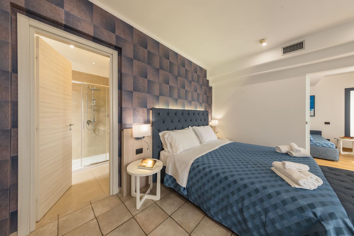 Une chambre d’hôtel moderne avec un grand lit, une petite table de chevet, un espace douche et une vue sur la chambre voisine.