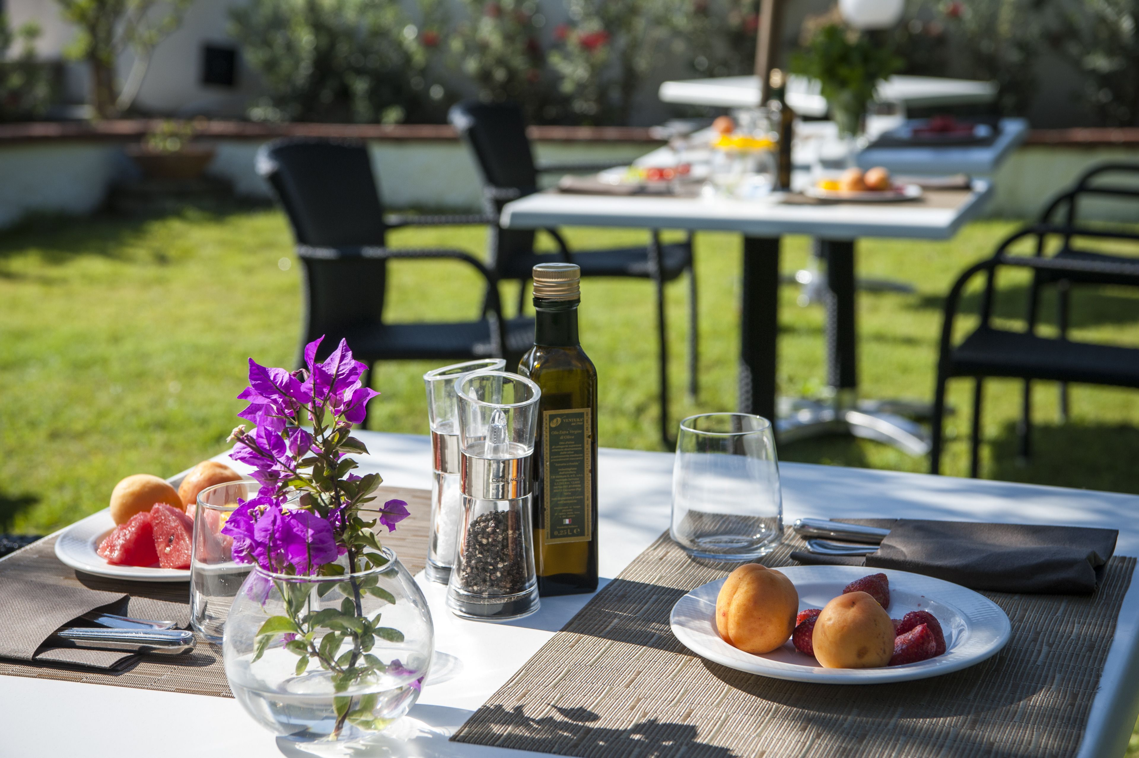 Une table de jardin dressée pour un repas en plein air, avec des assiettes remplies de nourriture, une bouteille d’huile d’olive et un vase contenant des fleurs violettes.
