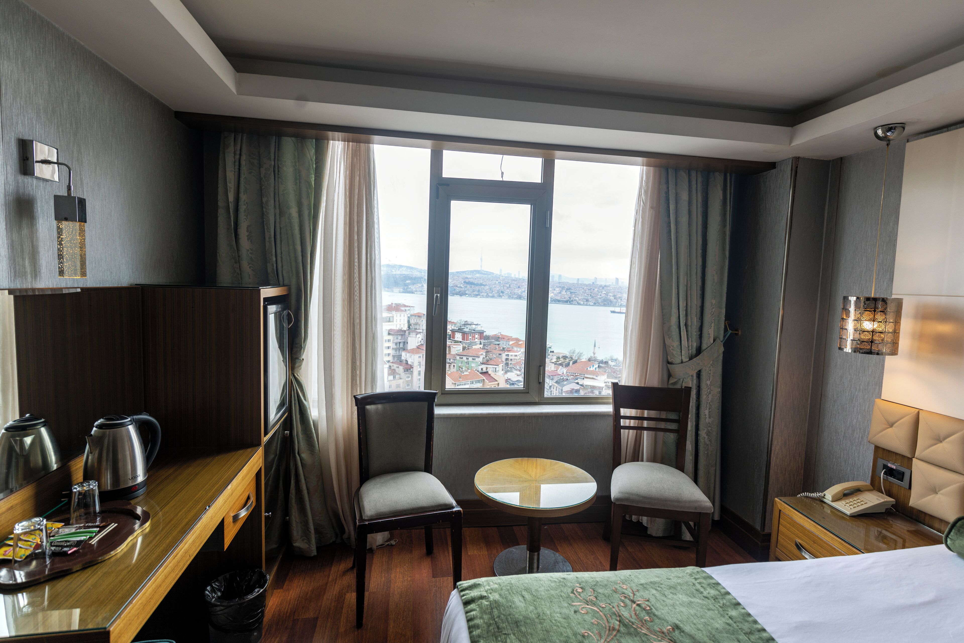 A hotel room with a bed, a desk with a kettle, a chair, a small table, and a view of a cityscape through a window.