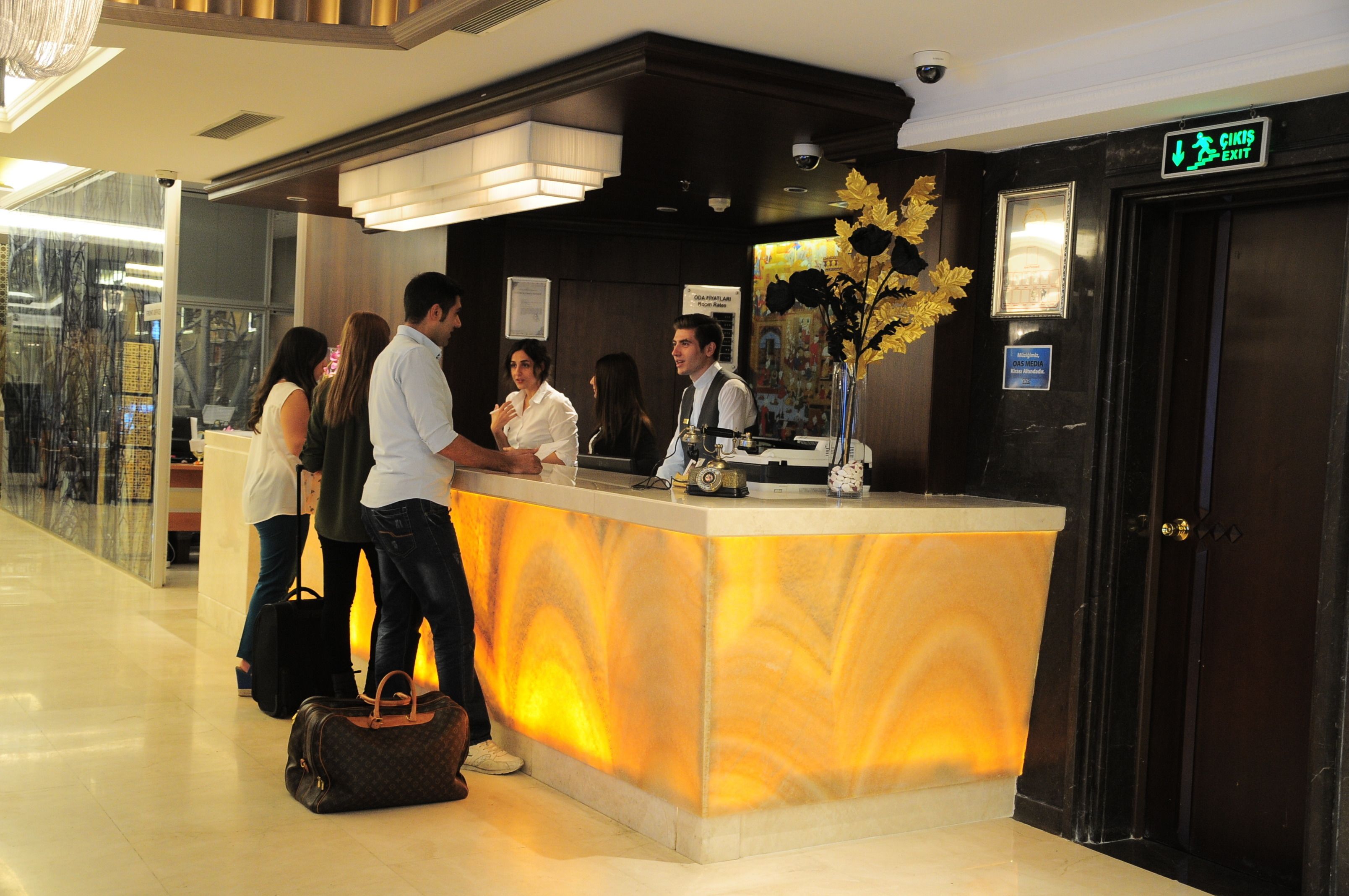 A hotel reception desk with a marble counter, a receptionist, and guests conversing.
