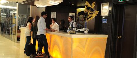 A hotel reception desk with a marble counter, a receptionist, and guests conversing.