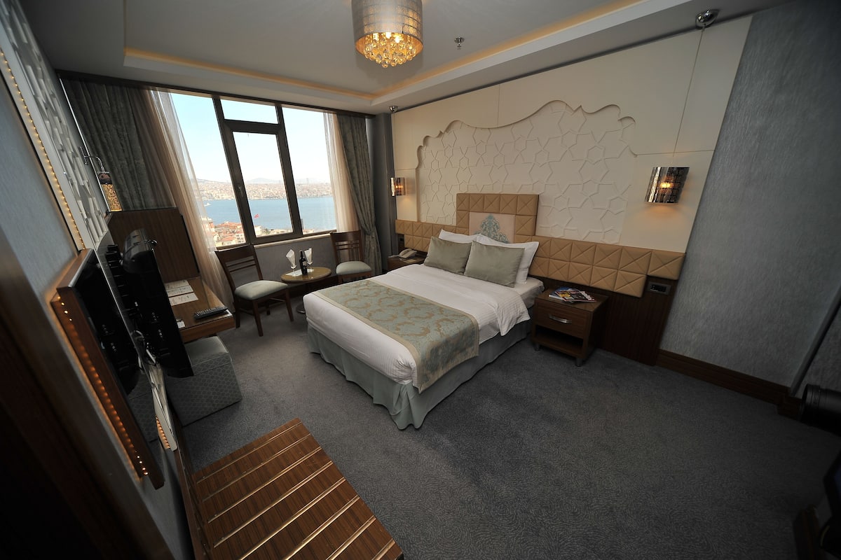 A hotel room with a large bed, a desk with a chair, a nightstand, a lamp, and a view of the city through the window.