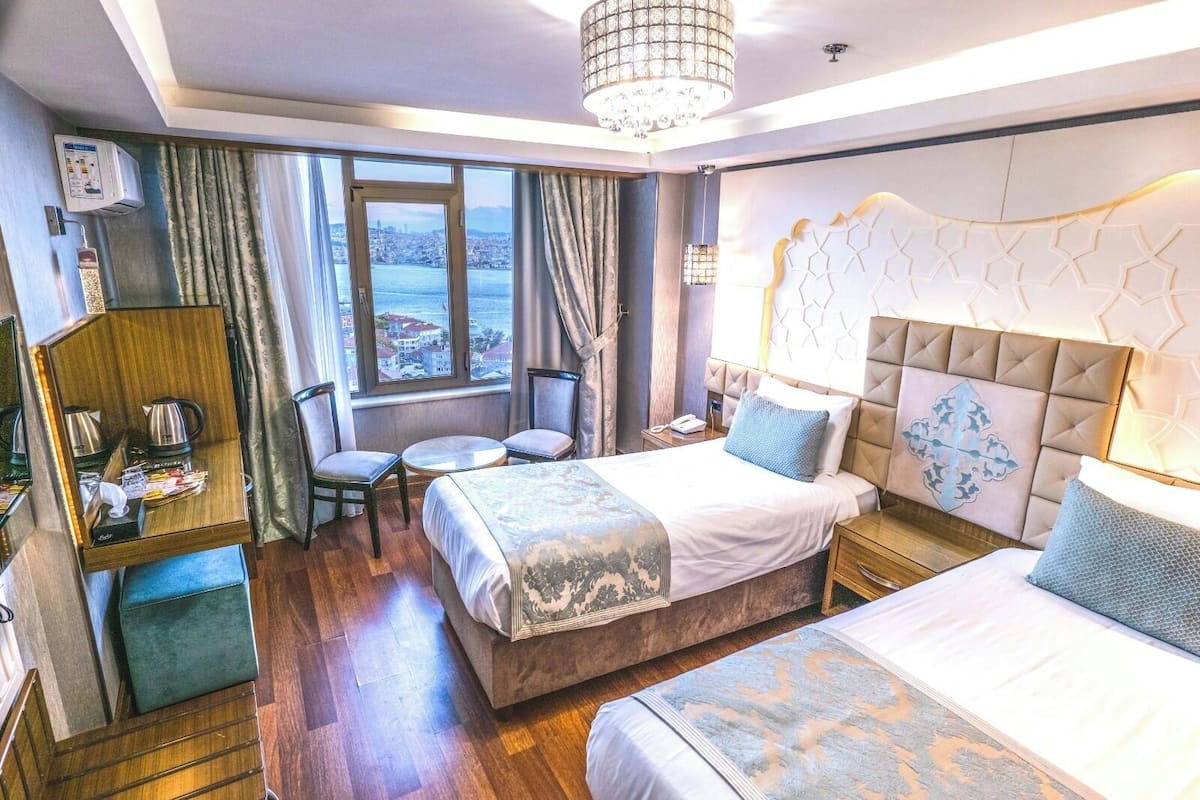 A hotel room with a large bed, a smaller bed, a desk with a coffee maker, a chair, and a view of the city through the window.