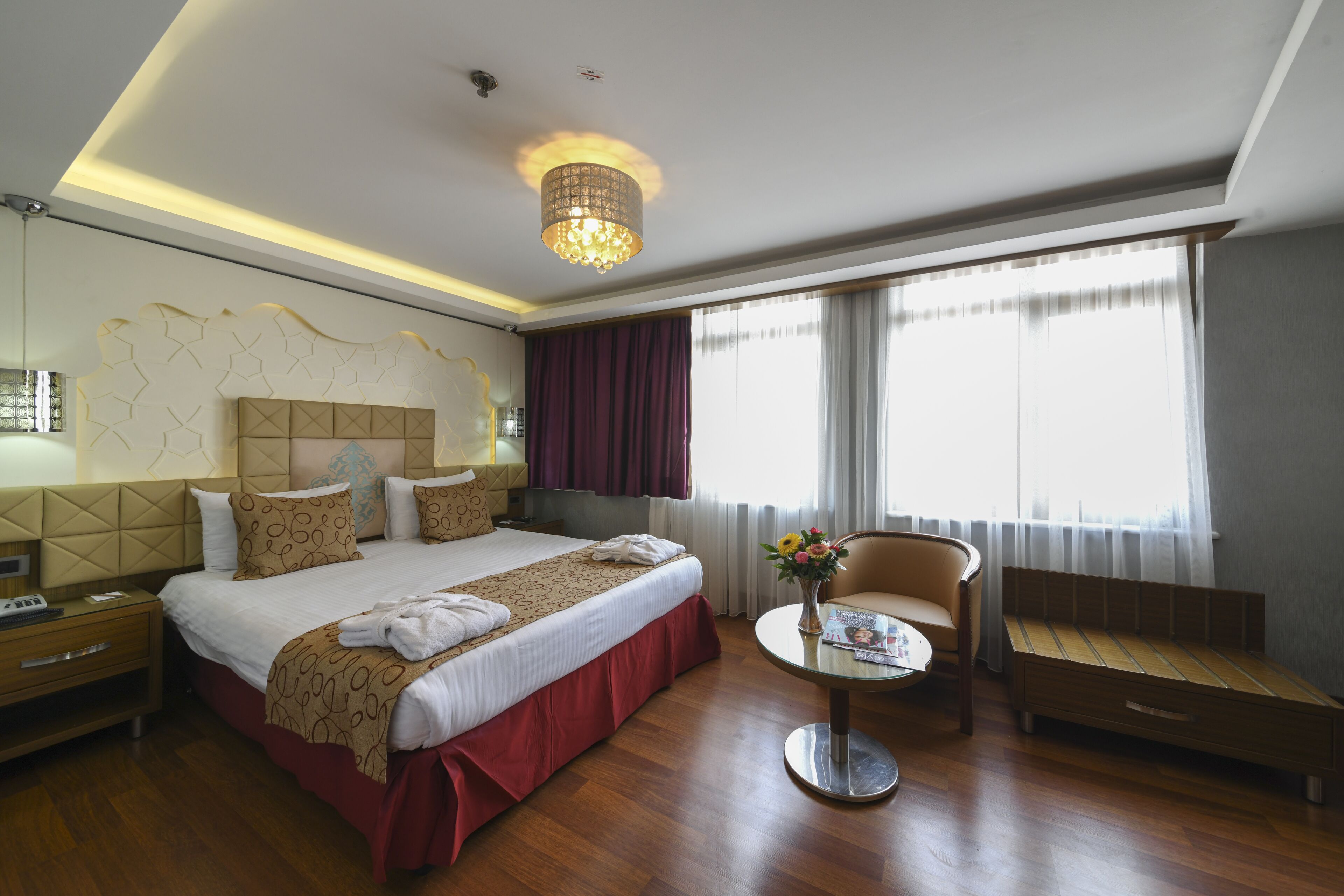 A hotel room with a large bed, a bedside table with a vase of flowers, a chair, a small table, and a window with curtains.