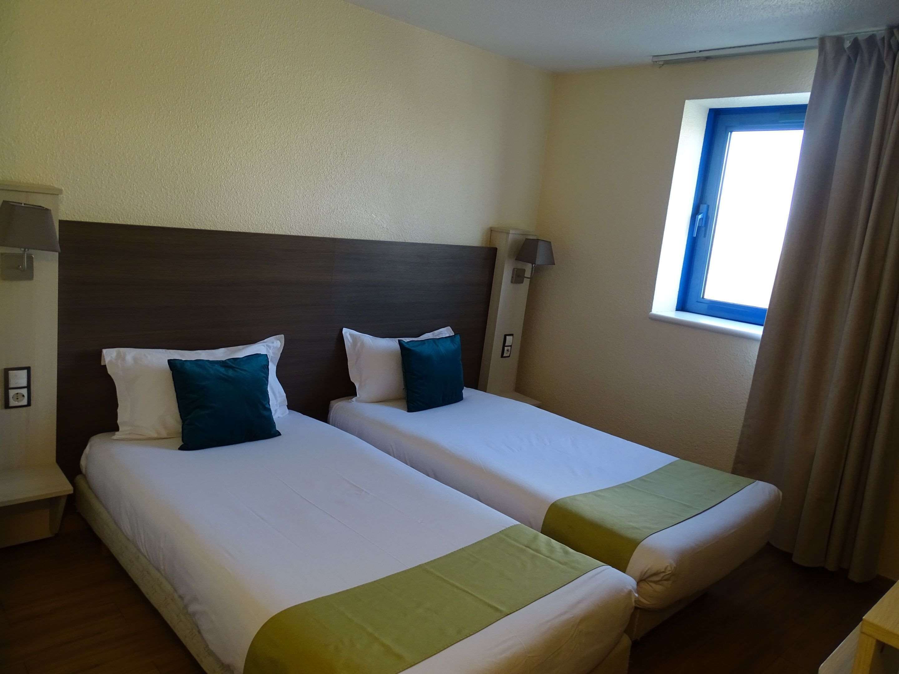 A hotel room with two beds, each with white linens and green bedspreads. There is a window with curtains, a nightstand with a lamp, and a wall-mounted light fixture.