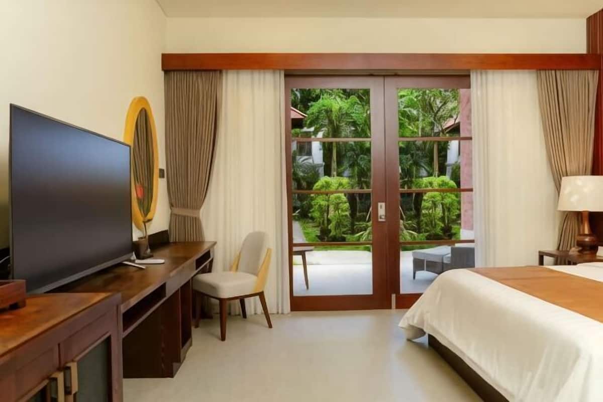 A hotel room with a TV, a desk, a chair, a bed, and a sliding glass door leading to a balcony with greenery.