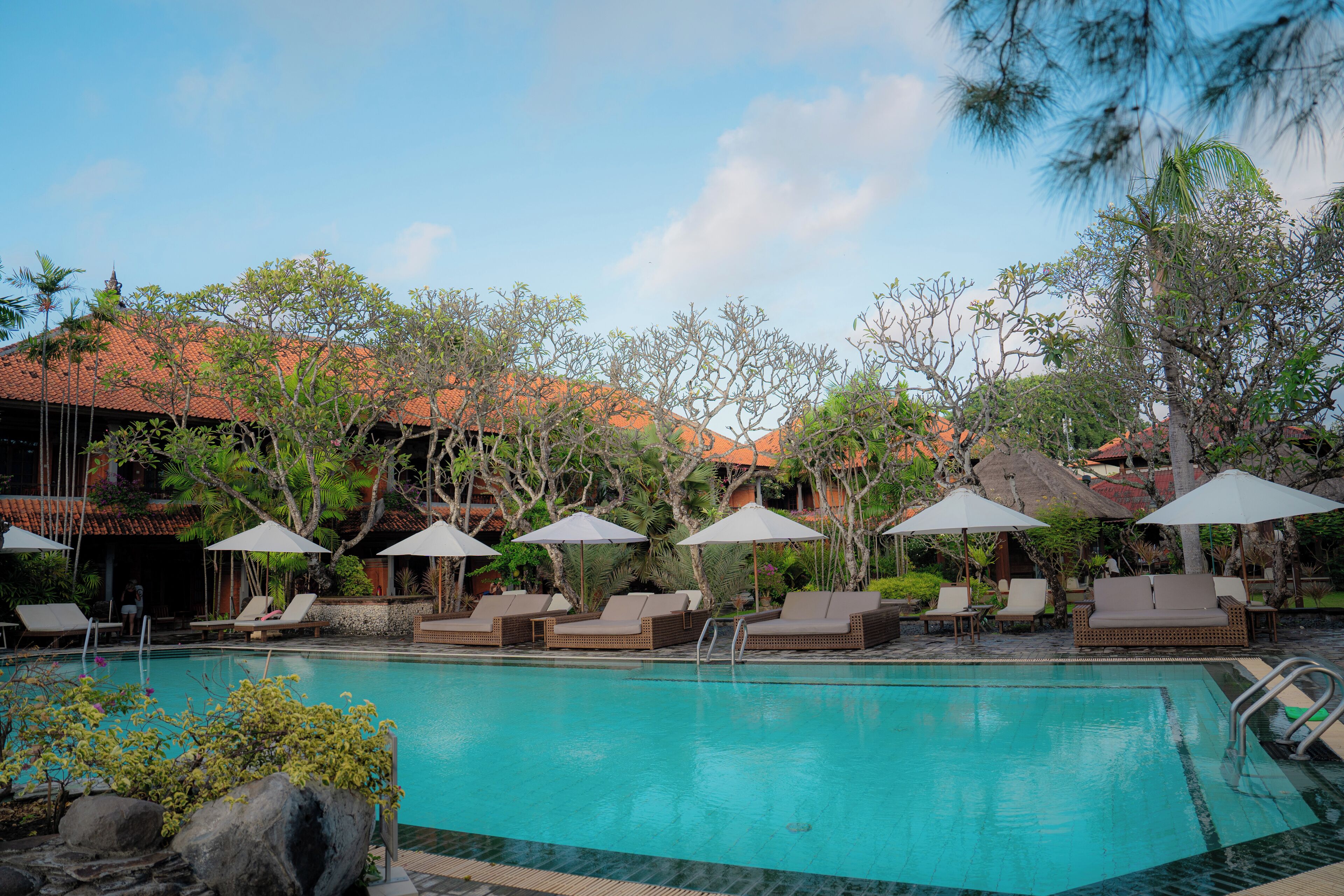 A pool area with lounge chairs and umbrellas, surrounded by trees and buildings with red roofs.