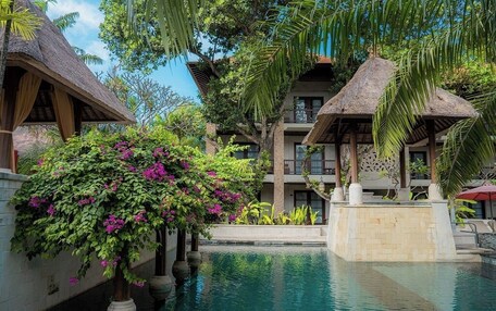 A pool area with a thatched-roof structure, lush greenery, and a multi-story building in the background.