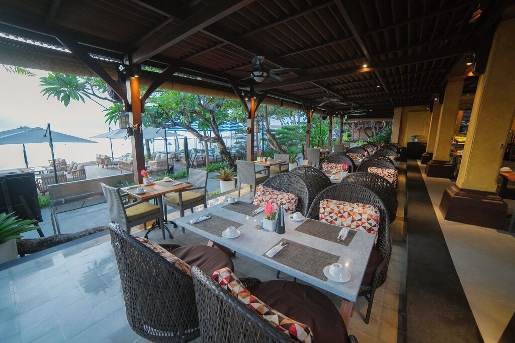 An outdoor restaurant with wicker furniture and tables, surrounded by greenery and overlooking a body of water.