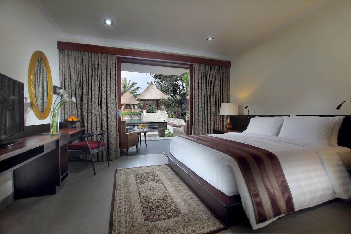 A hotel room with a bed, desk, chair, and a view of an outdoor area with umbrellas and plants.