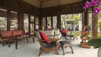 Un espacio interior amplio con mobiliario de madera, incluyendo un sofá, sillas y una mesa de centro. Se observa una recepción al fondo.