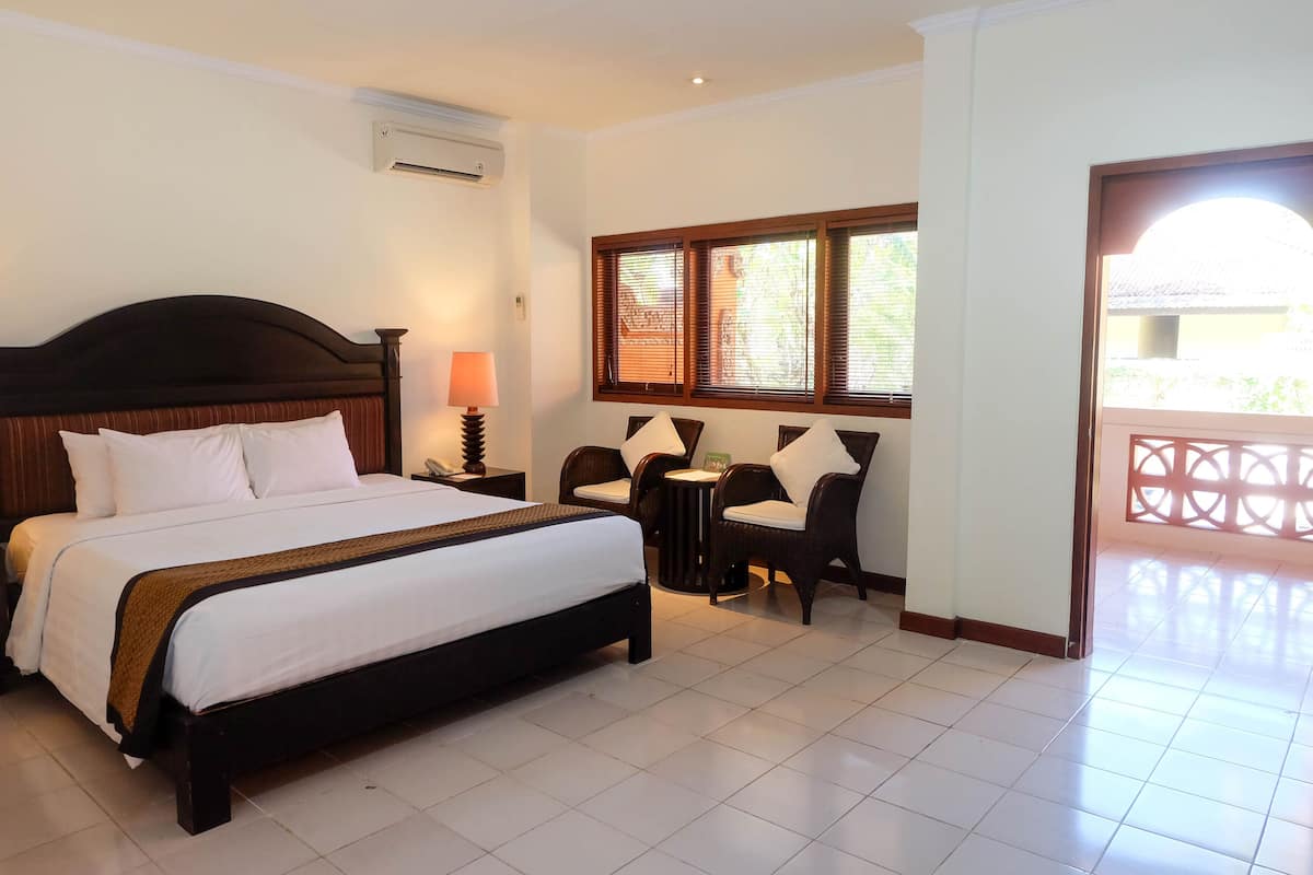 A hotel room with a large bed, a bedside lamp, a small table with chairs, and a window with blinds.