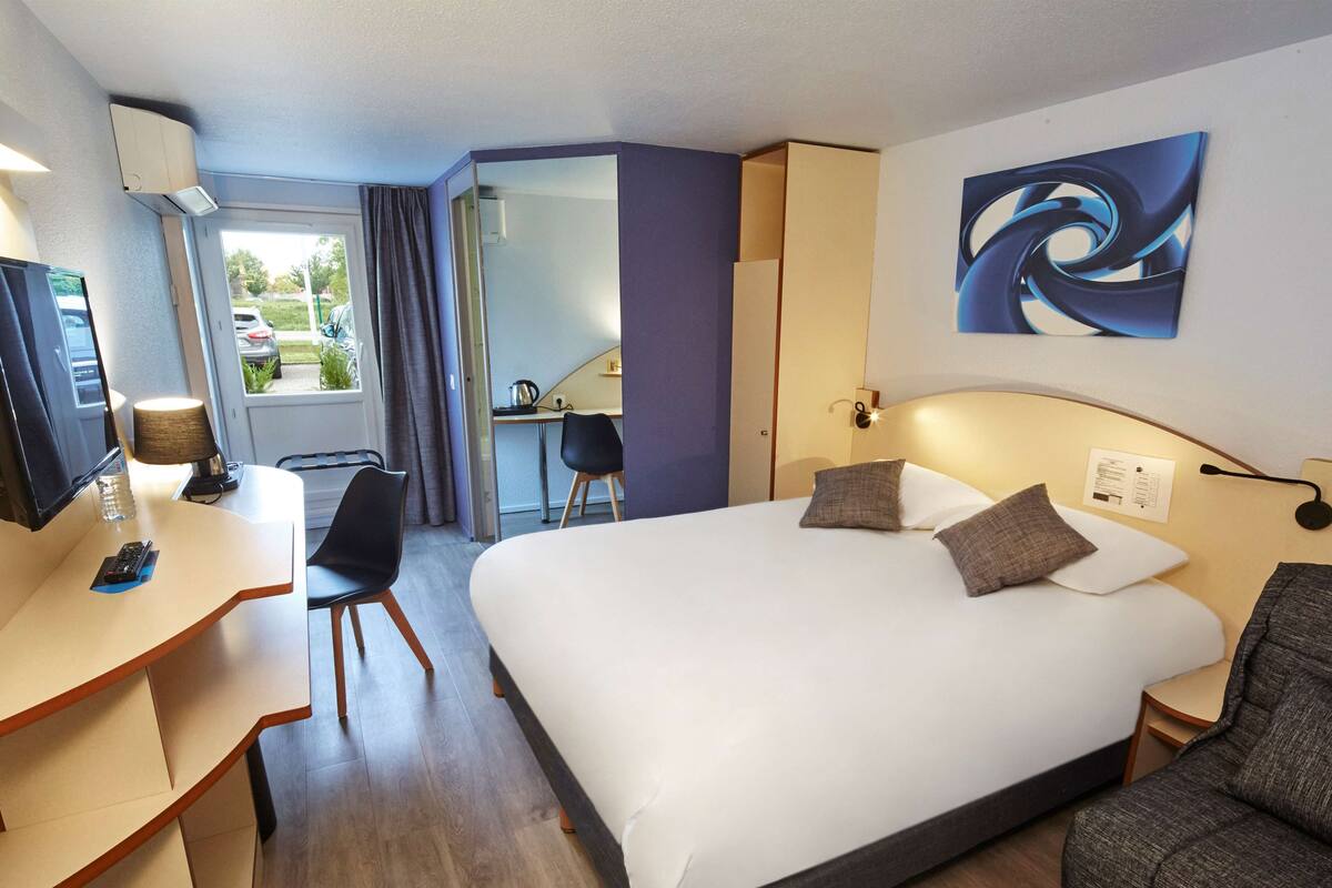 Quarto de hotel com cama, escrivaninha, cadeira, televisão e janela.