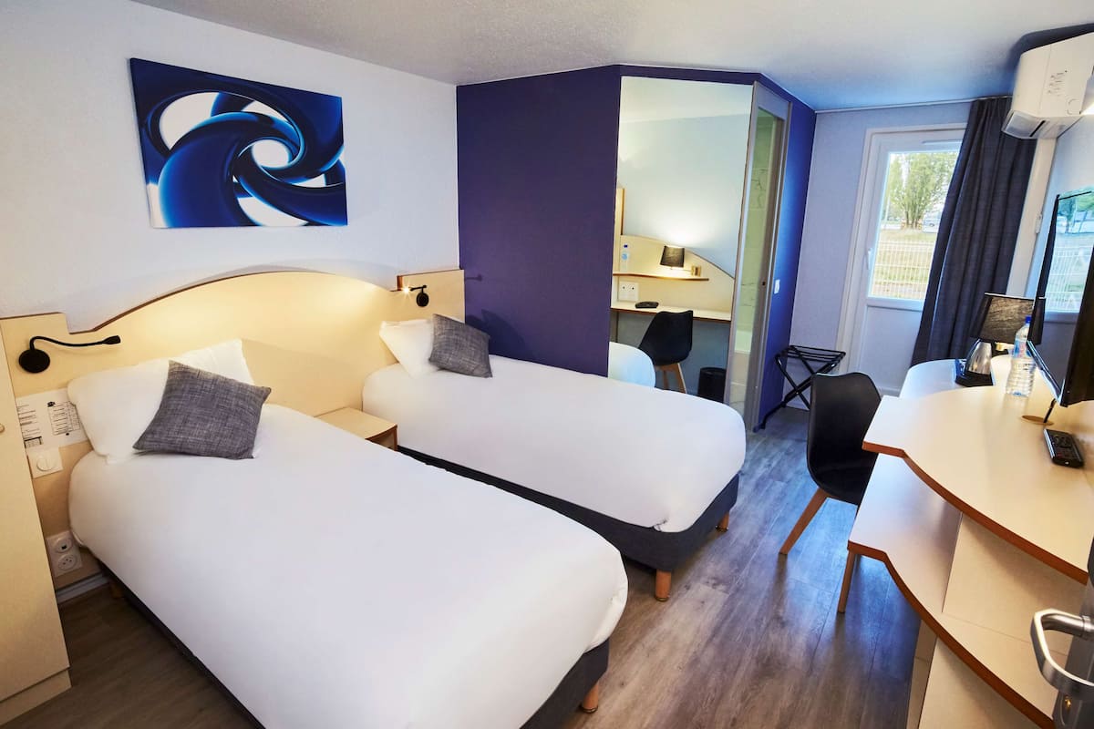 Quarto de hotel com duas camas de solteiro, uma escrivaninha, uma cadeira, um espelho e uma janela com cortinas.