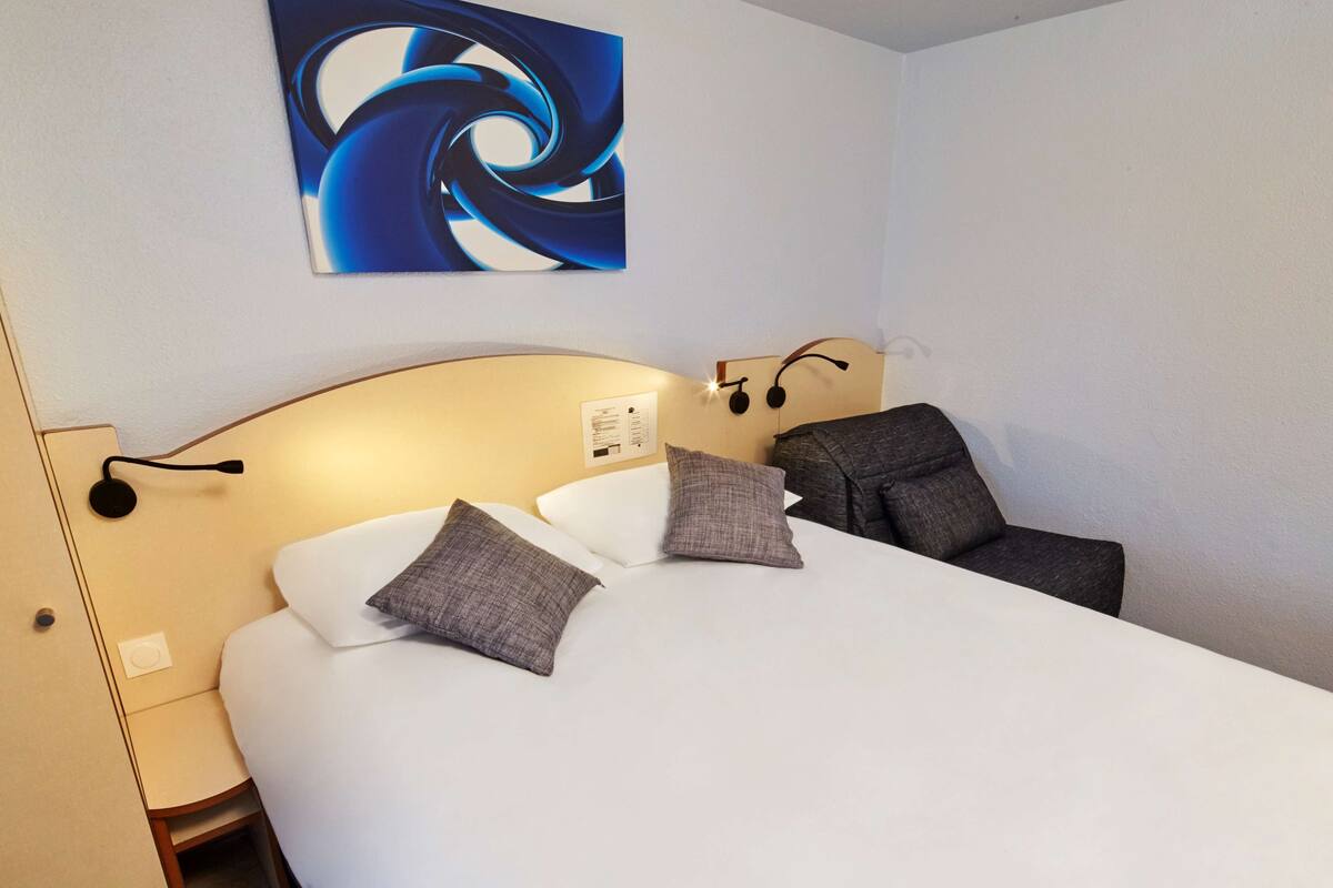 Quarto de hotel com cama, duas almofadas, cabeceira com iluminação e mesa de cabeceira com abajur, além de um quadro pendurado na parede.