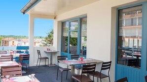 An outdoor dining area with tables and chairs, large windows, and a view of buildings and trees.