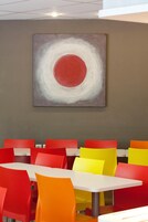 Un comedor con sillas rojas y amarillas, una mesa blanca y un cuadro abstracto grande en la pared.