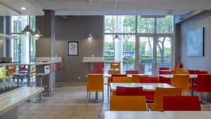 A modern cafeteria with red and yellow chairs, white tables, and large windows providing natural light. - Cyan Hotel Roissy Villepinte Parc Des Expositions (Villepinte)