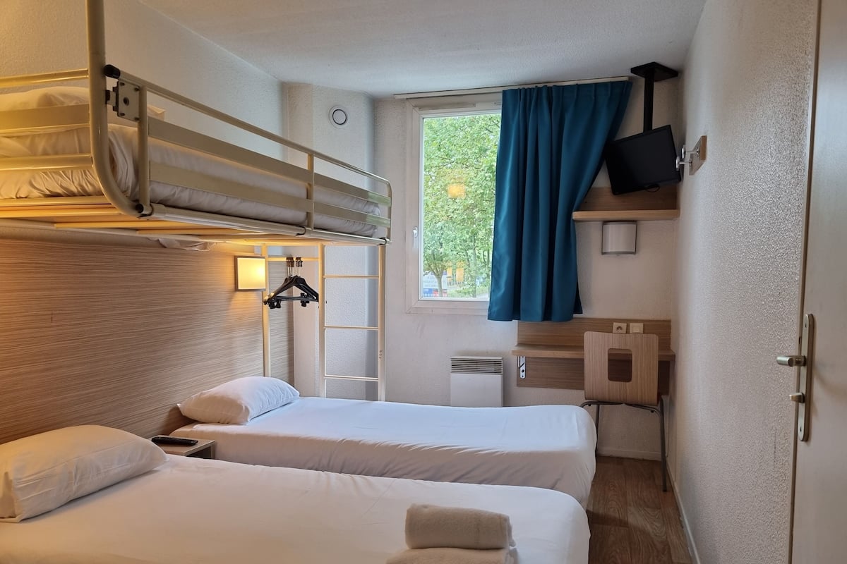 Une chambre d’hôtel compacte comprenant deux lits, un lit superposé, une télévision et une fenêtre avec des rideaux.