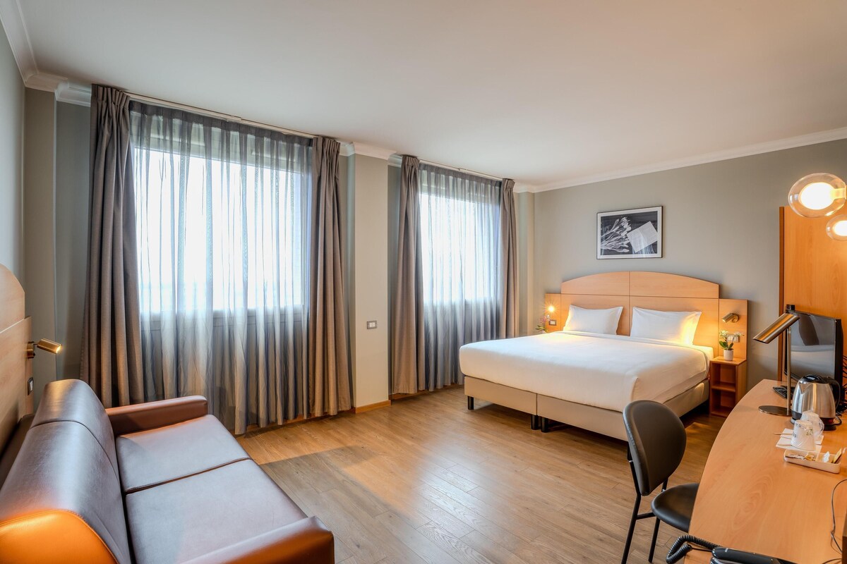 A hotel room with a bed, a desk, a chair, a sofa, and curtains.
