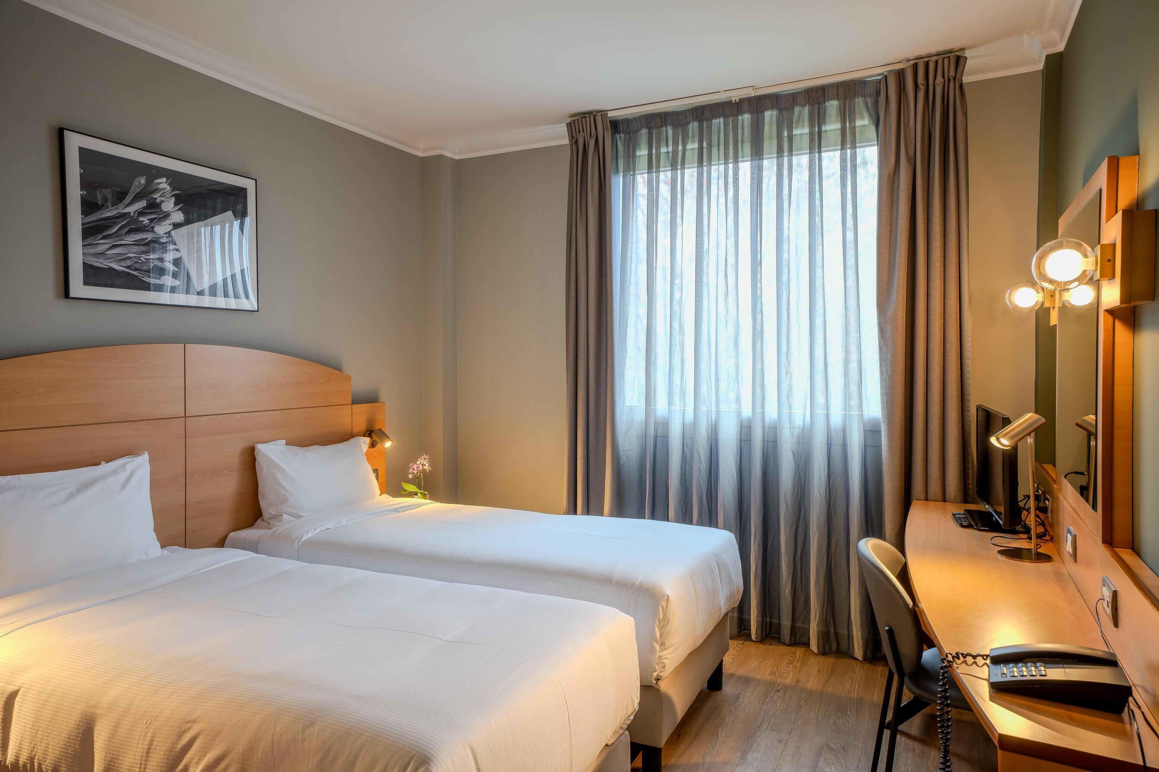 A hotel room with two beds, a desk with a telephone, a chair, a desk lamp, and a large window with curtains.